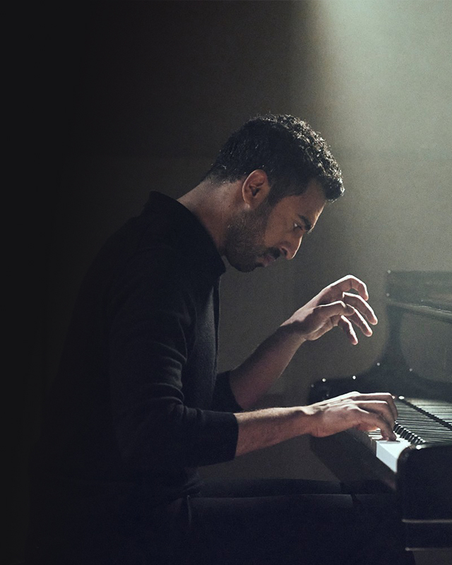 A man wearing a black jumper playing the piano under a spotlight.